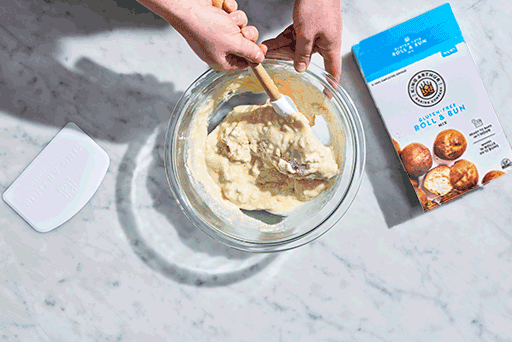 Mixing Gluten-Free Roll and Bun Mix in a clear bowl with a spatula.