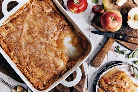 A square ceramic pan of Sugar-Crusted Apple Cobbler with some portioned into a small bowl and a few fresh apples to the side - select to zoom