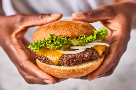Holding a hamburger that is loaded up with cheese, onion, and lettuce all between a gluten-free roll. - select to zoom