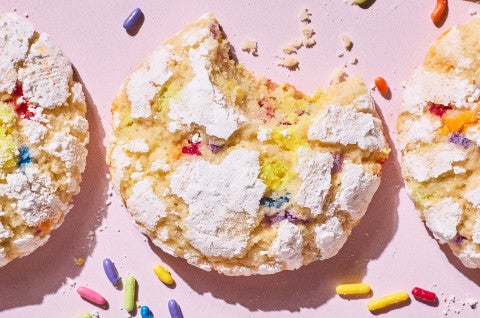 Sprinkle Crinkle Cookies on a light lavender background and a bite taken out of a cookie - select to zoom