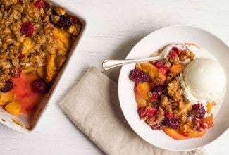 A pan of freshly baked, crumb-topped Summer Fruit Crisp next to a bowl of fruit crisp with a spoon
