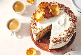 An overhead photo of a Hummingbird Cake with pineapple cream cheese frosting. A slice has been taken out of the cake and there are toasted nuts and a slice of dried pineapple on top of the cake. 