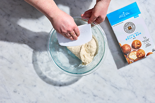 Folding Gluten-Free Roll and Bun Mix in a clear bowl with a bowl scraper..