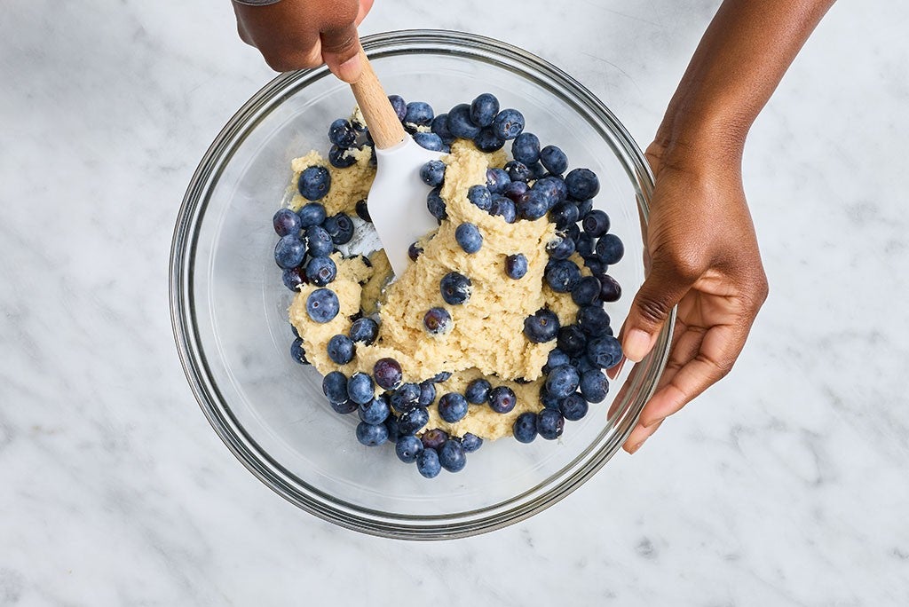 A person mixing Emancipation Breakfast Cake batter in a glass bowl with blueberries