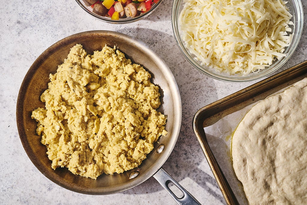 Scrambled eggs in a pan, shredded cheese in a bowl, and a pizza crust stretched onto a sheet tray all seen from above.