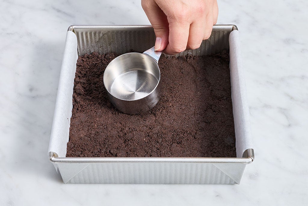 Patting the crumb crust of No-Bake Chocolate Cheesecake Bars with a measuring cup