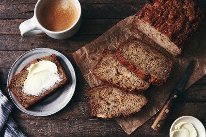 A loaf of Sourdough Banana Bread cut into slices, with one piece slathered in butter
