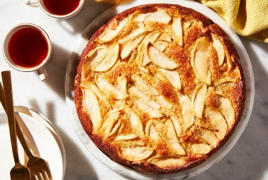 A freshly baked Mostly Apples Apple Cake next to two cups of tea and forks